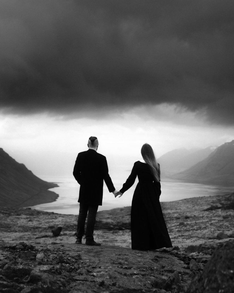 Daria Endresen and Hjalti Sveinsson, the two photographers at Ast og Hraun, holding hands and facing a fjord in Icelandic Westfjords.